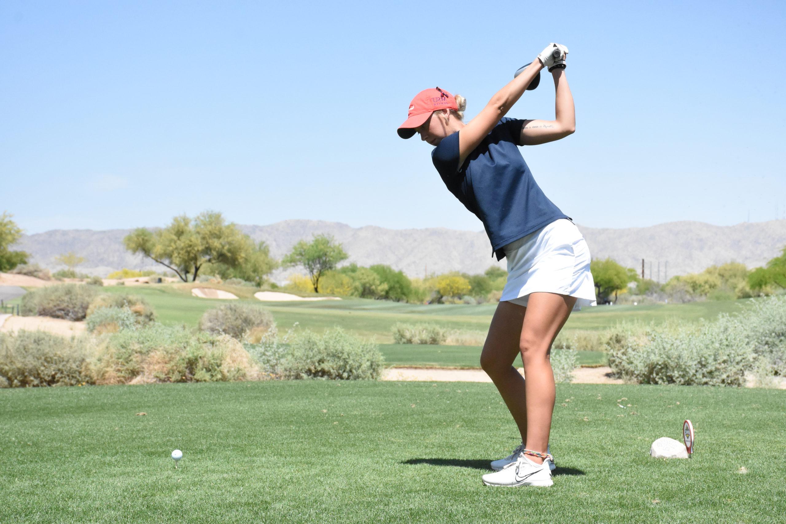 Golf athlete Courtney Lawler tees off at a desert golf course with a mountain range and trees in front of her.